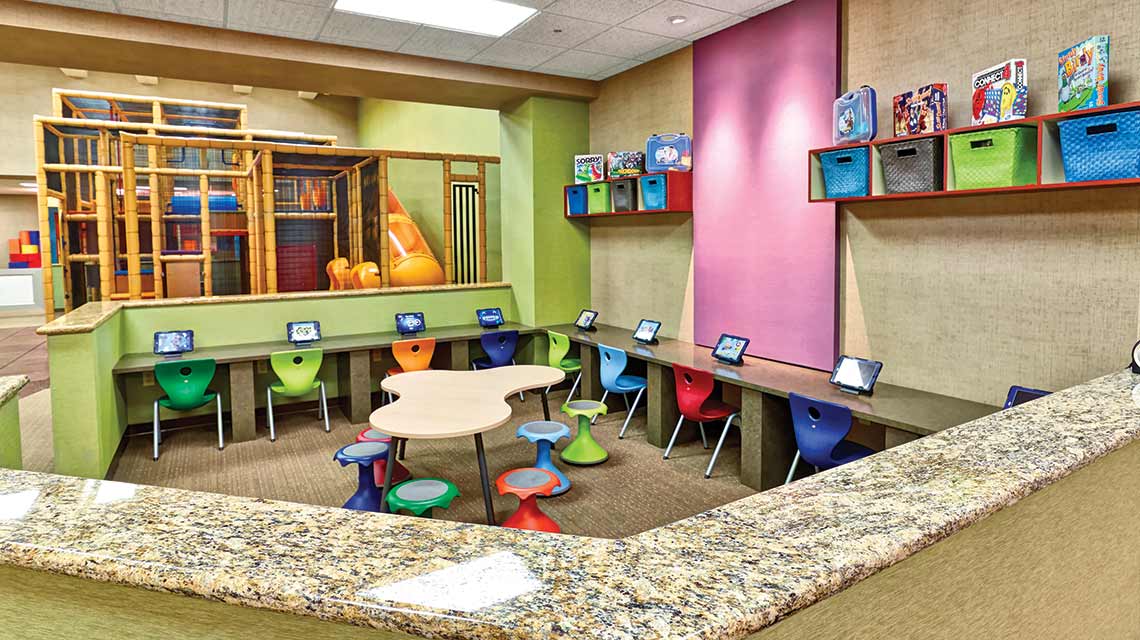 An aerial view of a children’s media room with a row of tablets set on a low marble countertop with small, colorful children’s chairs 