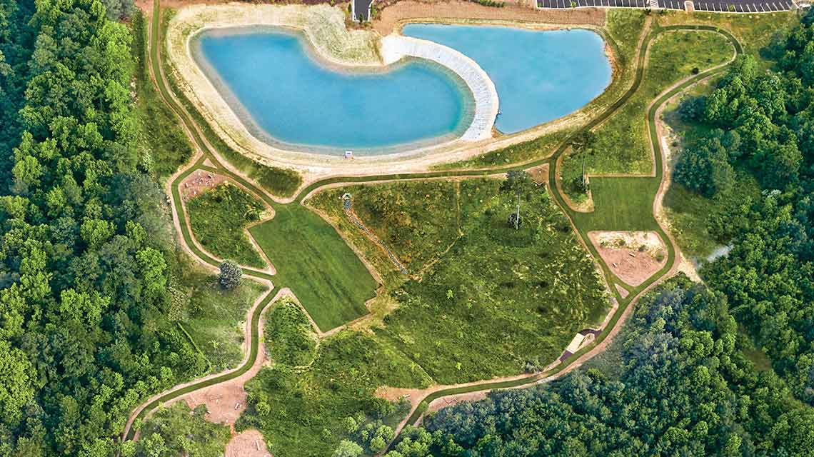 An aerial view of a crystal-clear blue outdoor pool surrounded by an outdoor fitness trail, grass and green trees 