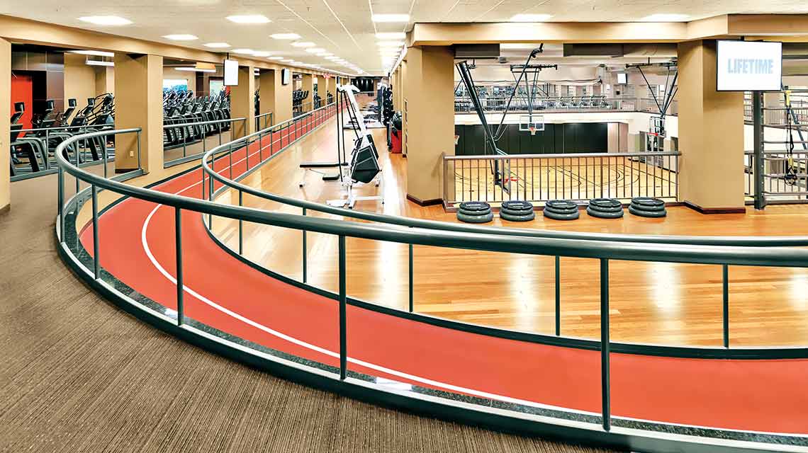 A wooden fitness floor circled by a red walking/running track with metal railings on both sides and cardio machines in the background