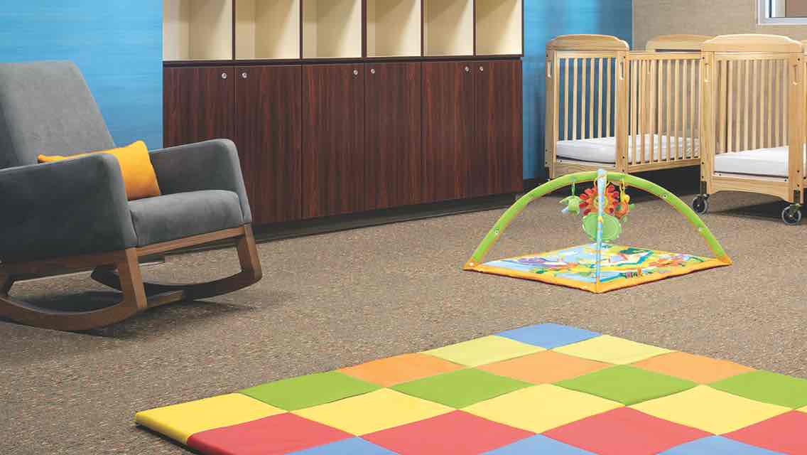 A carpeted infant room with a cushioned rocking chair, a row of wooden cabinets and cubbies, play floormat, an infant play mobile and two wooden cribs