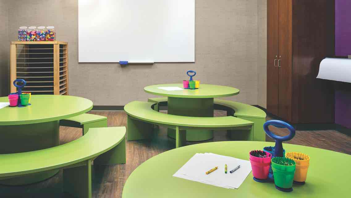 A children’s language arts studio equipped with three green tables surrounded by bench seating with containers of crayons and paper
