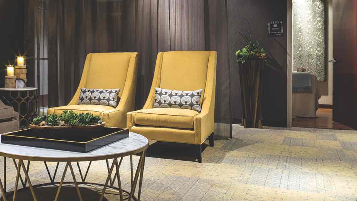 A luxe, candle-lit spa waiting area with cushioned yellow arm chairs, lush carpeting and green plants 