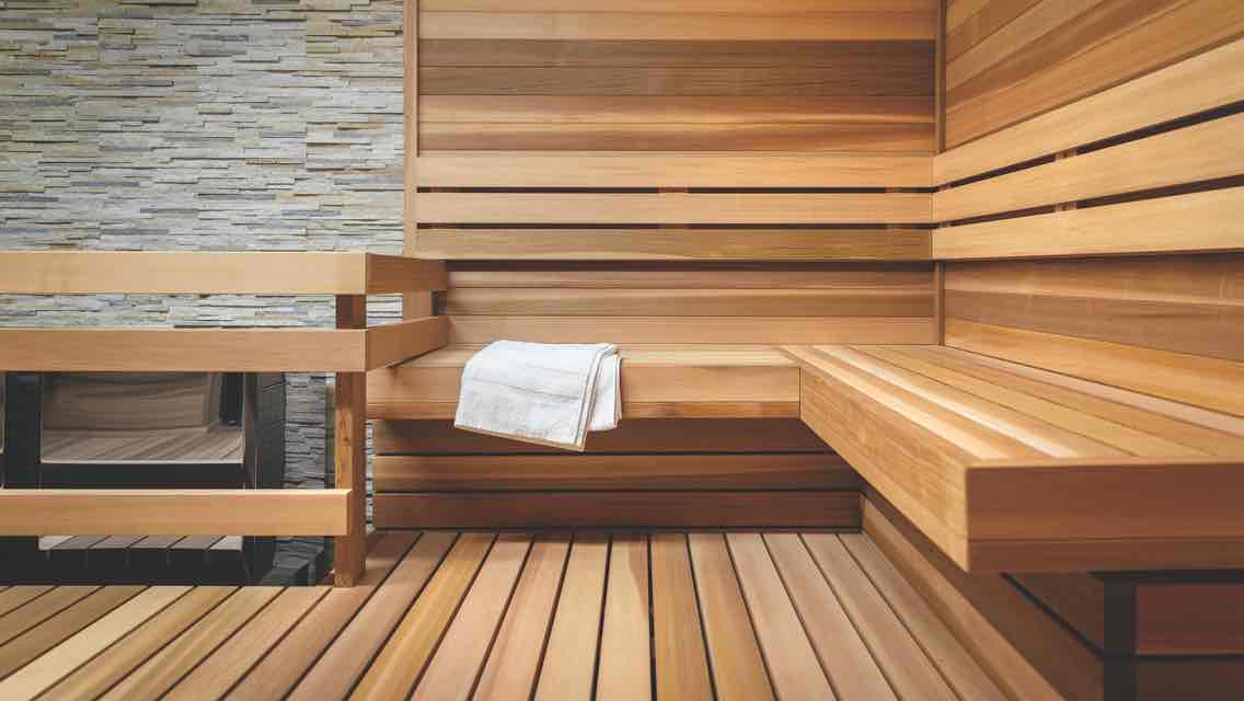 : A pristine sauna with tile walls, wood floors and a wood bench holding a crisp white towel 