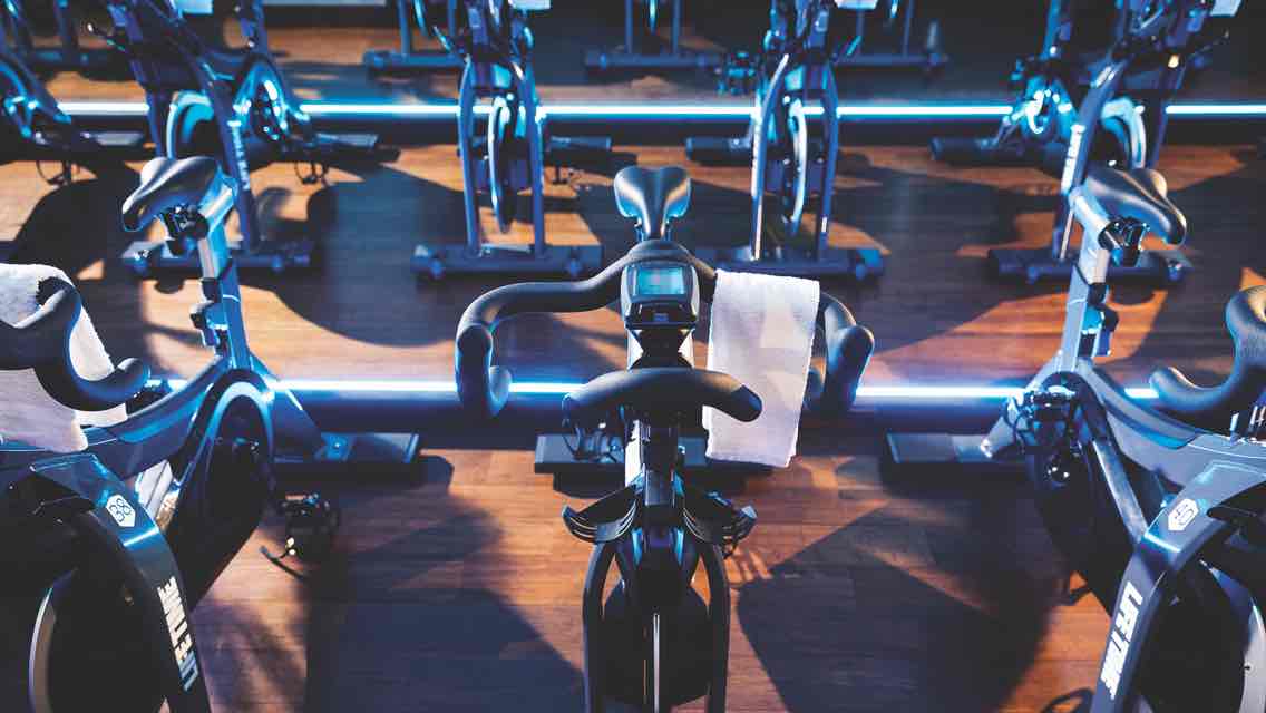 Rows of state-of-the-art stationary bikes face the instructor’s platform in a purple- and blue-lit cycle studio