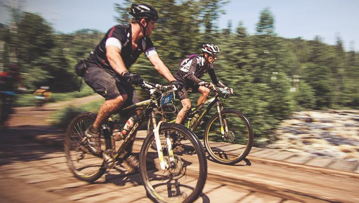 Two bikers in Life Time Cycle gear racing on a trail through a forested area