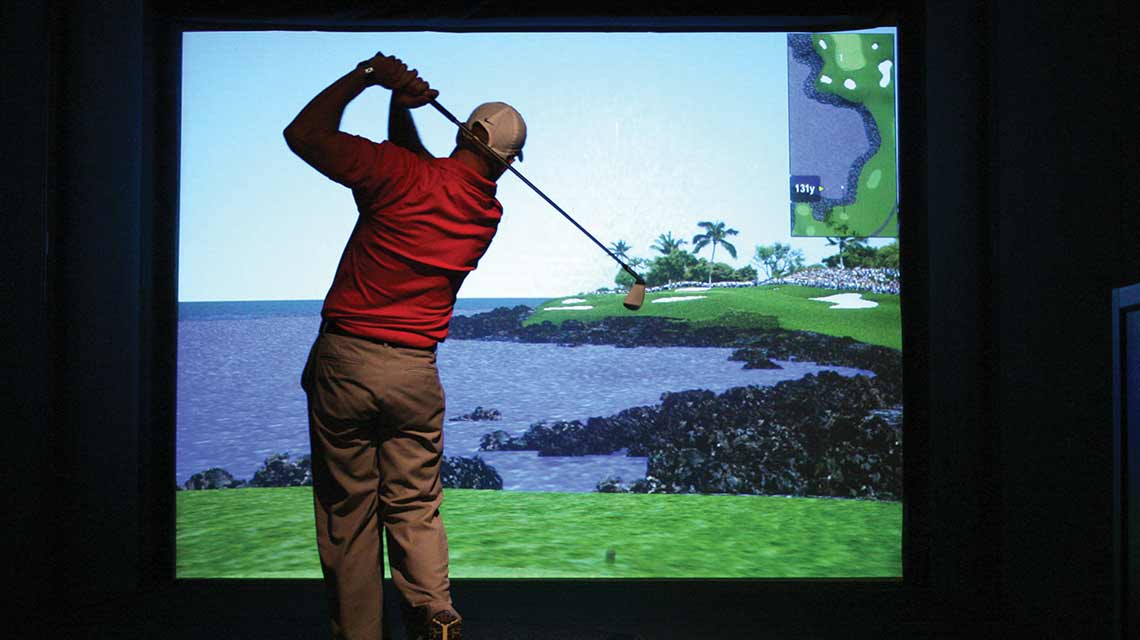 A man swings a golf club behind his back while overlooking a video simulation of grass, trees and water