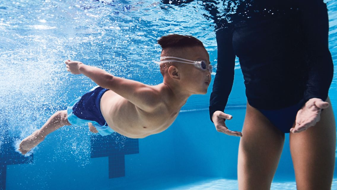 A woman guides a child wearing swim goggles through the pool water