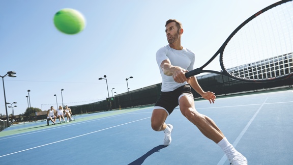 A man in a white tennis shirt and black shorts takes a one-handed backhand swing with a tennis racquet at a lofted tennis ball on an outdoor tennis court.