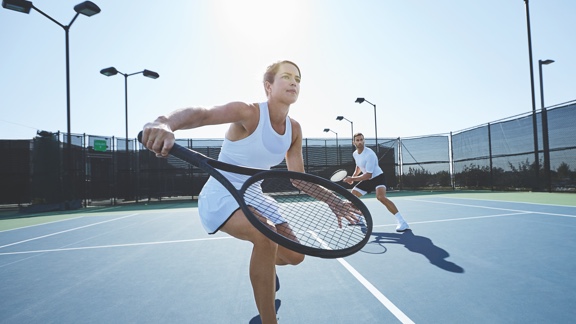 A woman in a white tennis outfit takes a one-handed backhand swing with a tennis racquet on an outdoor tennis court
